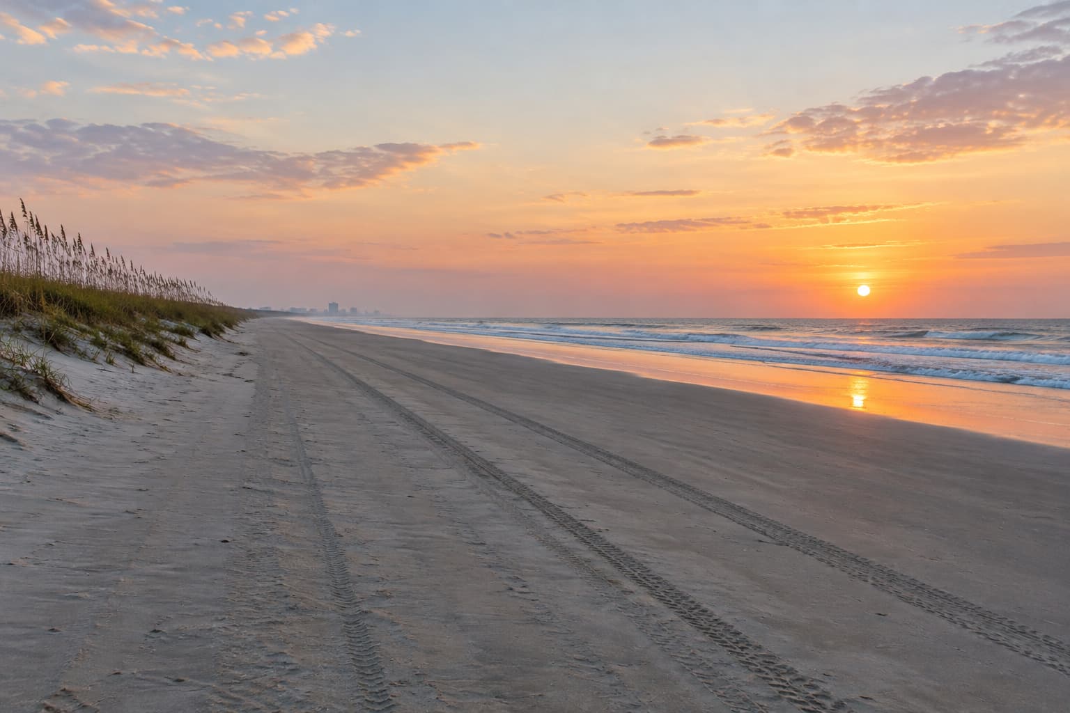 A quiet Daytona Beach shoreline at sunrise, with sea oats along the dunes and tire tracks in the sand leading toward the ocean